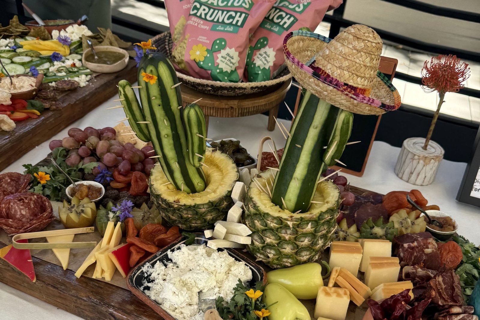 Charcuterie board with a cactus sculpted from cucumbers and toothpicks planted in a hollowed pineapple, beside bags of Cactus Crunch tortilla chips