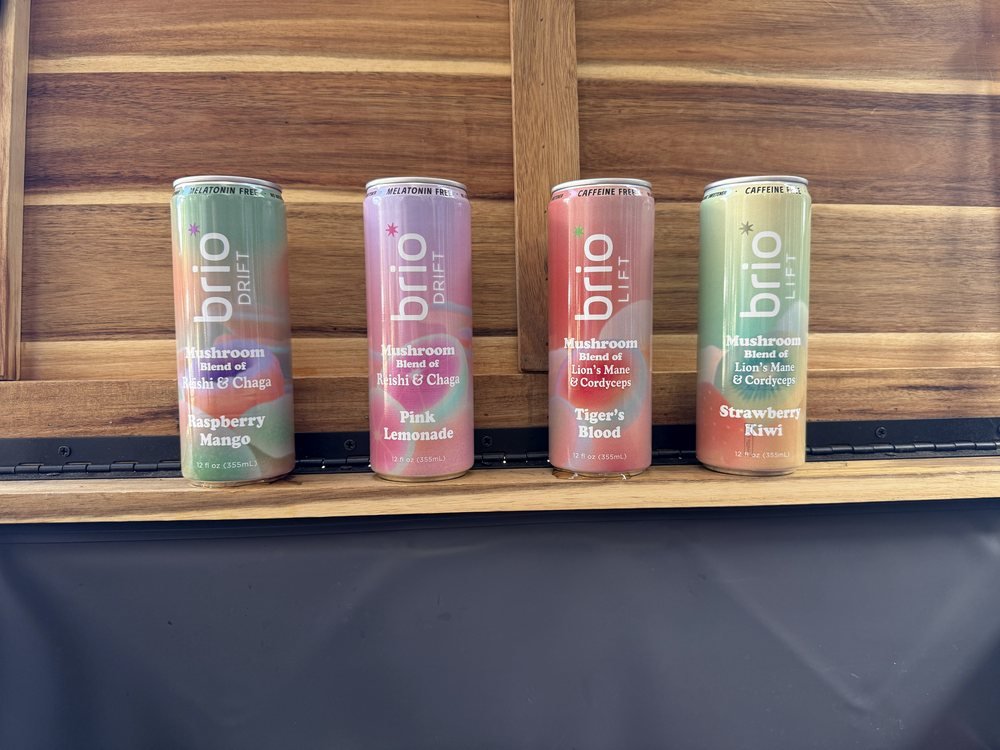 Four cans of Brio mushroom beverages lined up on a wooden surface: Drift Raspberry Mango, Drift Pink Lemonade, Lift Tiger's Blood and Lift Strawberry Kiwi