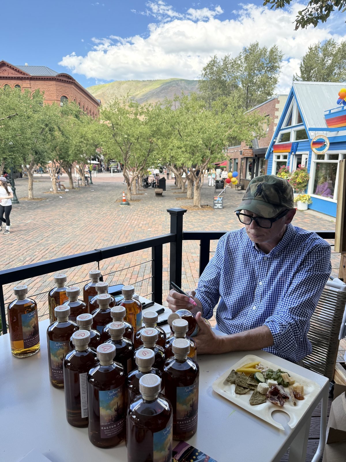 Artist Simon Bull drawing on bottles of Cactus Añejo at a table on the Bosq patio, with Aspen's brick pedestrian mall behind him