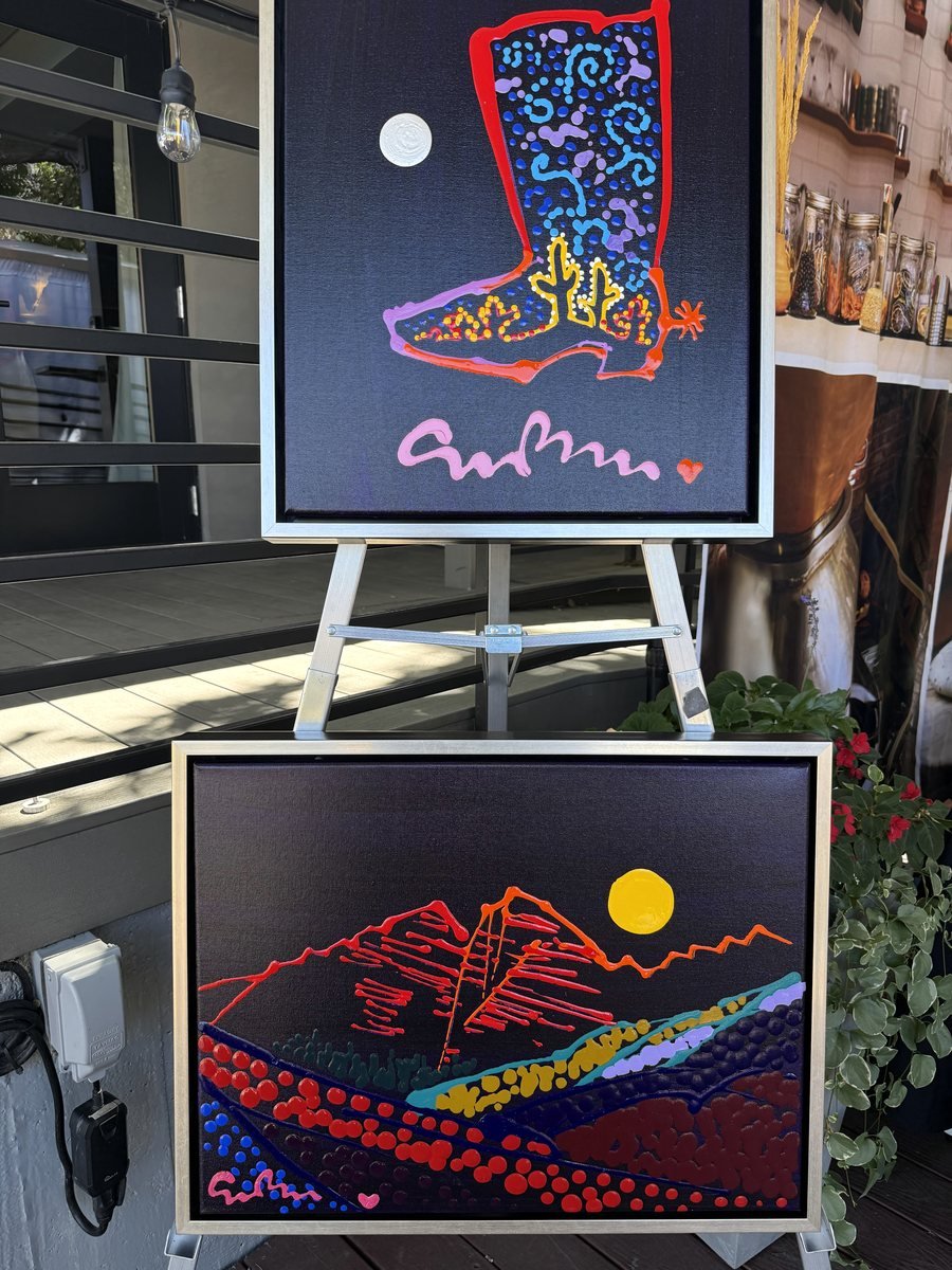 Two framed Simon Bull paintings on easels: a cowboy boot filled with cactus forms, and a mountain landscape under a yellow sun
