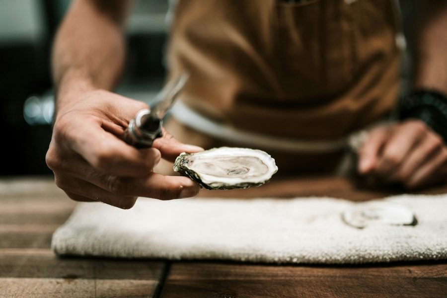 A hand opening an oyster with a shucking knife over a folded towel on a wooden counter