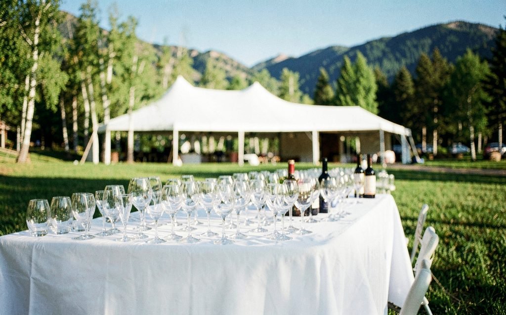 A long white-linen table set with wine glasses in a mountain-town park in summer, a white tent behind
