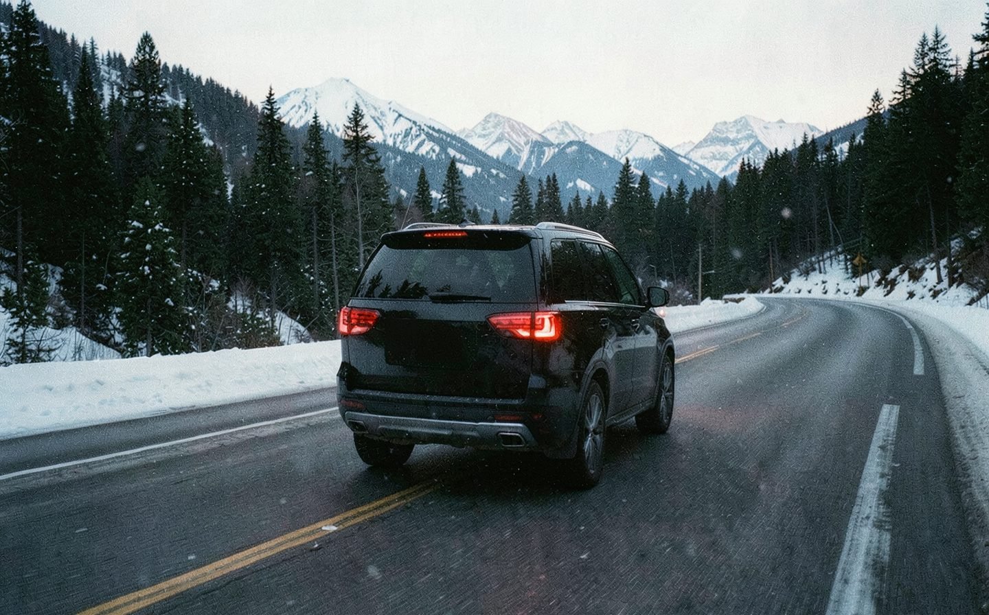 A black SUV driving along a snowy mountain highway at dusk, tail lights glowing