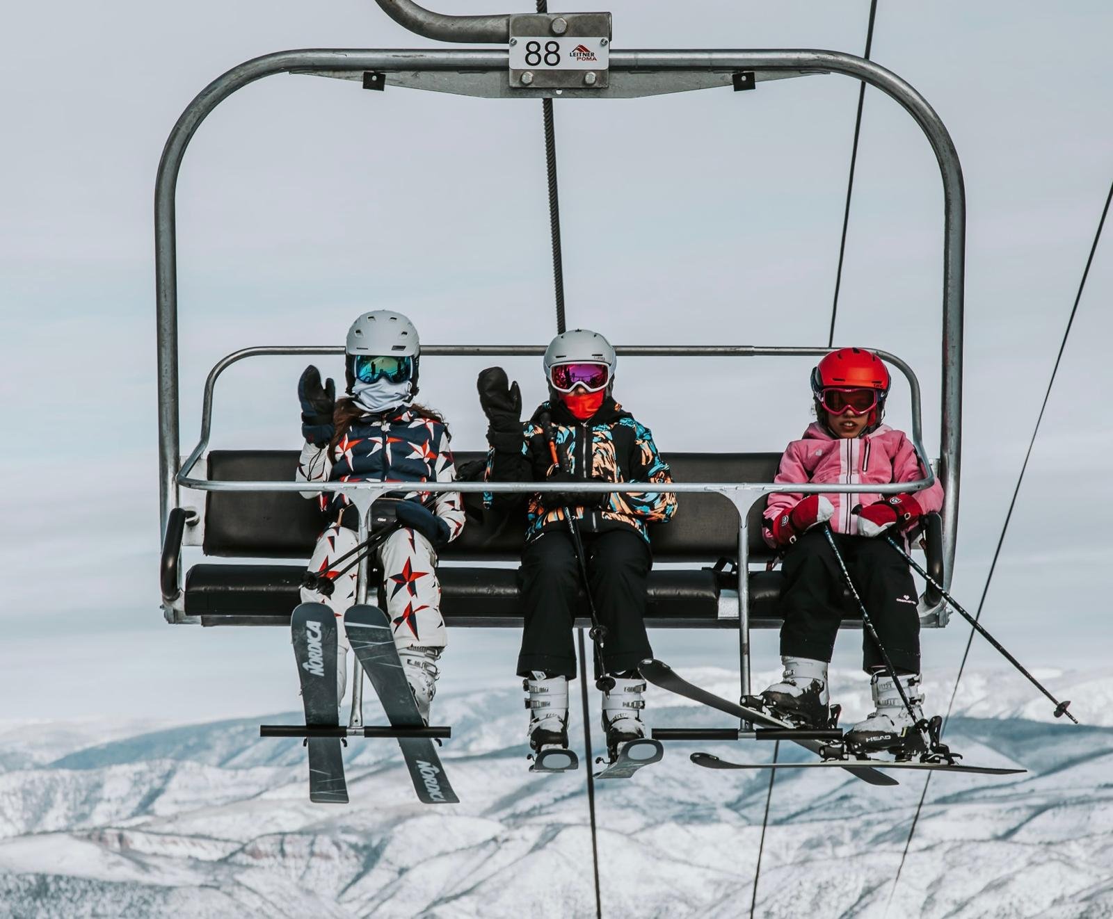 Three children on a ski chairlift in Aspen, waving with snow-covered mountains in the background
